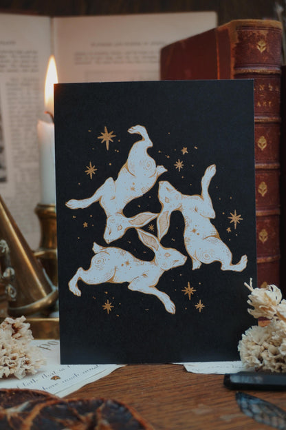 Postcard with a triple hare design in white and gold on a black background, resting on a wooden table with books, dried flowers and candles