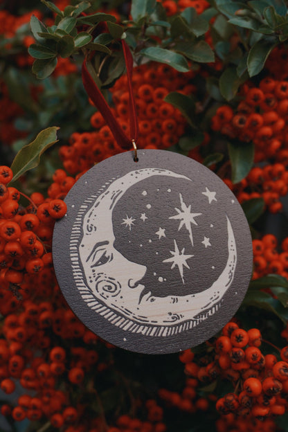Circular wooden hanging ornament with a celestial moon and stars design, resting on a red berry bush