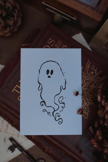 Swirly black outline ghost drawing on white card, resting on a vintage book surrounded by decorative items like dried flowers, vintage paper and a key in the background