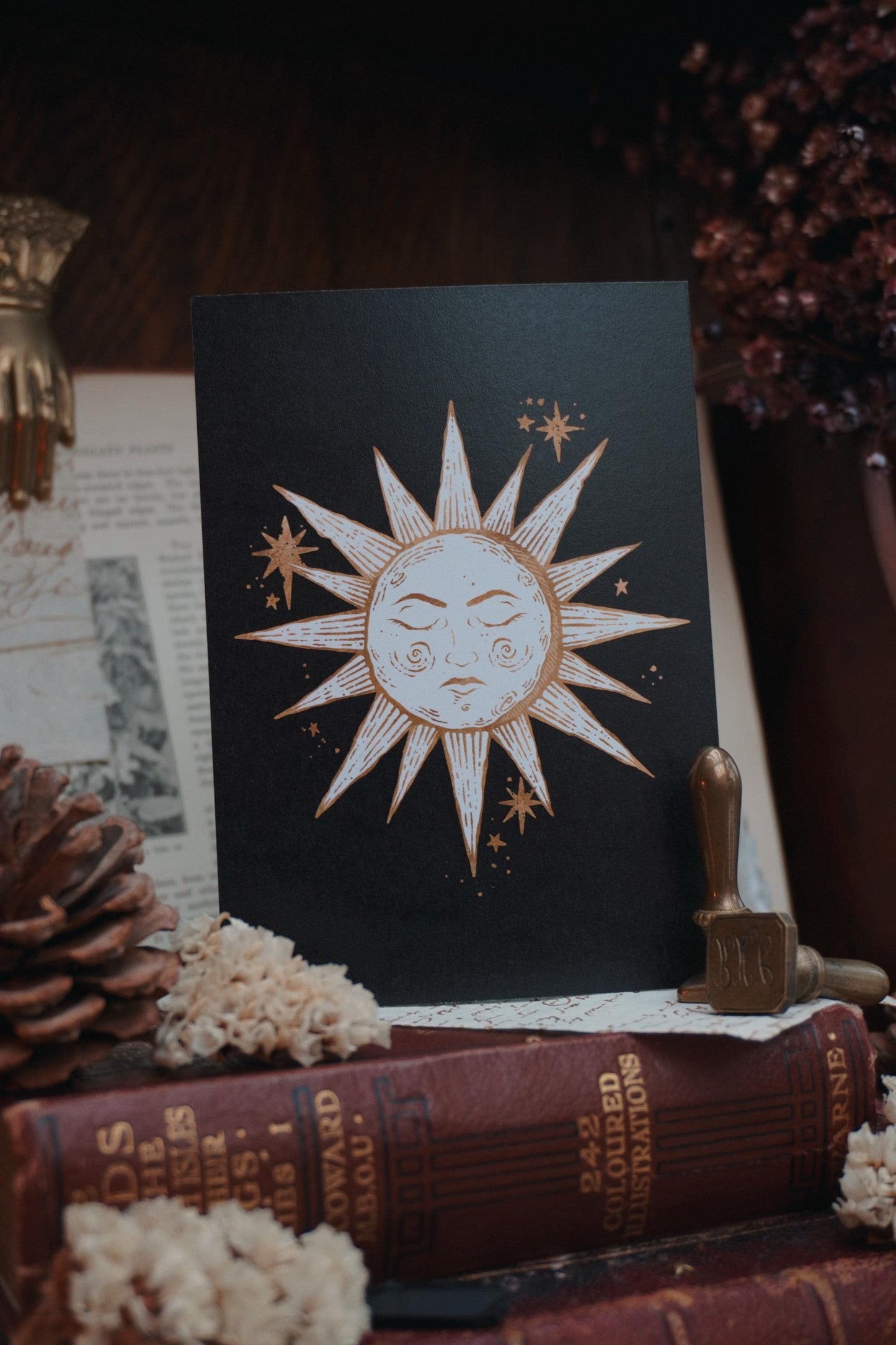 Postcard with a sun design in white and gold on a black background, resting on a wooden table with books and dried flowers