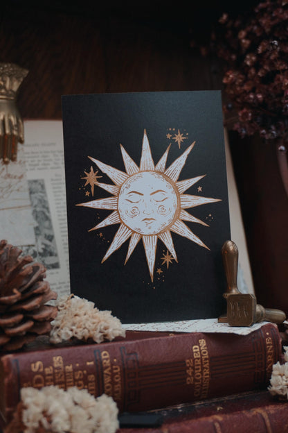 Postcard with a sun design in white and gold on a black background, resting on a wooden table with books and dried flowers