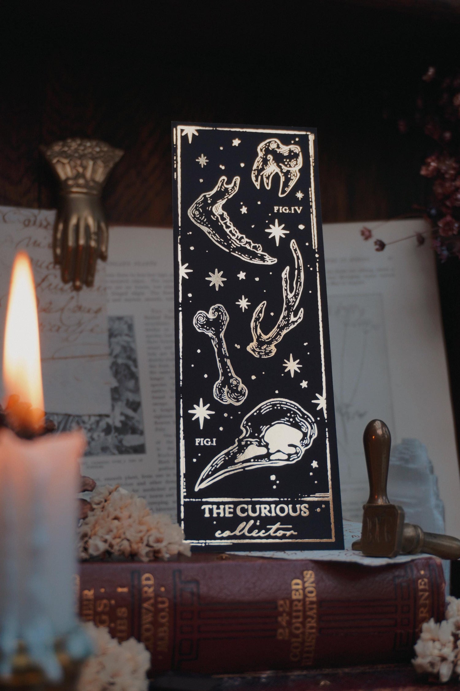 Black bookmark with Gold various bones design resting on vintage books surrounded by decorative elements including a lit taper candle, dried flowers and  brass wax seal stamps